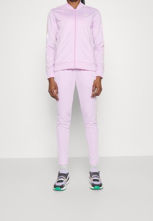 Person trägt eine hellrosa Reißverschlussjacke und passende Hose, weiße Knöchelsocken und mehrfarbige Sneakers, steht vor einem einfarbigen Hintergrund.