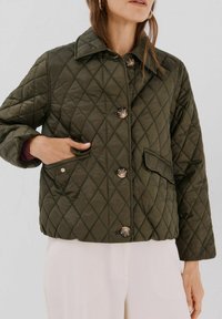 Giacca trapuntata verde oliva con colletto, dotata di grandi bottoni, tasche laterali e una vestibilità ampia. Tessuto texturizzato con cuciture a rombi.