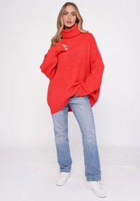 Przesadnie duży sweter dzianinowy w jasnym czerwonym kolorze z wysokim kołnierzem, z szerokimi rękawami i rozciętymi bokami, w połączeniu z jasnoniebieskimi dżinsami i białymi sneakerami.