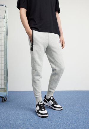 Personne portant un pantalon de survêtement Nike gris clair avec des poches zippées noires et des baskets Nike noir et blanc, debout sur un tapis bleu.