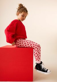 Suéter acolchado rojo, leggings rosas con patrones de corazones rojos y zapatillas negras de caña alta con suelas blancas. Niño sentado sobre un cubo rojo.