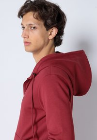 Un hoodie bordeaux en tissu doux, doté d'une capuche avec cordon de serrage, d'une poche kangourou et d'un petit logo sur la poitrine, créant un look décontracté.