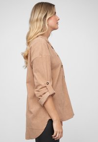 Lysebrun kordfløyelsskjorte med knapper, oppbrettede ermer, buet kant og teksturert stoff. Svart leggings bæres under.