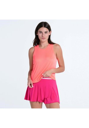 Junge Frau, die ein korallenfarbenes ärmelloses Oberteil und einen leuchtend pinken, plissierten Rock trägt, steht vor einem schlichten, hellen Hintergrund.