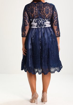 Mujer vistiendo un vestido azul marino de encaje hasta la rodilla con mangas tres cuartos y tacones altos nude, vista desde atrás.