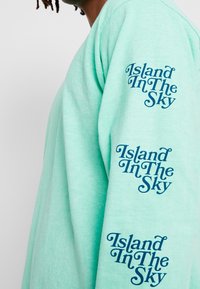 Minttu vihreä pitkähihaisa paita pehmeällä tekstuurilla. Ominaisuuksina navy sinisessä painatuksessa "Island In The Sky" molemmilla hihoilla.