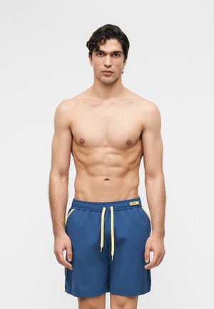 Muž bez trička s krátkymi tmavými kučeravými vlasmi, ktorý má na sebe modré plavkové šortky so žltými sťahovacími šnúrkami a bočnými vreckami, stojaci pred jednoliatym pozadím.