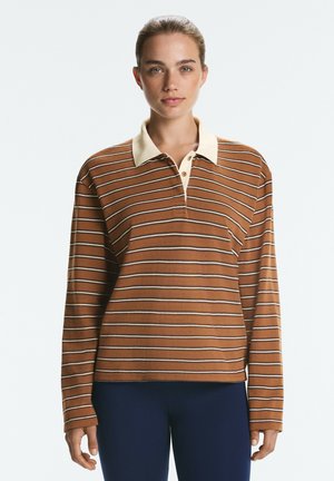 Femme aux cheveux attachés portant un polo marron à manches longues rayé avec un col crème et un pantalon sombre, debout devant un fond blanc.