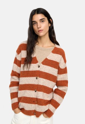 Kobieta o długich ciemnych włosach, ubrana w pomarańczowo-beżowy pasiasty sweter na guziki, na beżowym topie, stojąca z rękami w kieszeniach białych spodni.