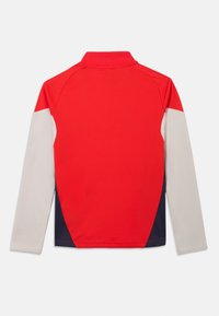 Κόκκινο πουκάμισο με μακριά μανίκια και ψηλό γιακά· διαθέτει λευκές και ναυτικές λεπτομέρειες στα μανίκια και στις πλευρές. Λείο, ελαφρύ ύφασμα.