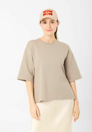 Femme portant un t-shirt oversize beige et une casquette assortie avec le texte « WOW MOM », debout devant un fond blanc uni.