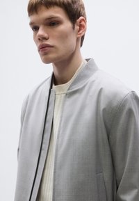 Világosszürke bomber dzseki texturált felülettel, álló gallérral, cipzáras lezárással és oldalzsebekkel. Krémszínű bordázott pulóver fölött viselve.