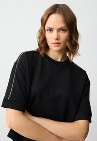 Czarny t-shirt z krótkim rękawem o okrągłym dekolcie z metalicznym akcentem wzdłuż szwu na ramieniu. Gładka faktura i luźny krój.