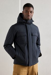 Casaco acolchoado azul-marinho com capuz, apresentando dois grandes bolsos frontais, um fecho éclair à frente e uma textura suave. Usado sobre calças bege.