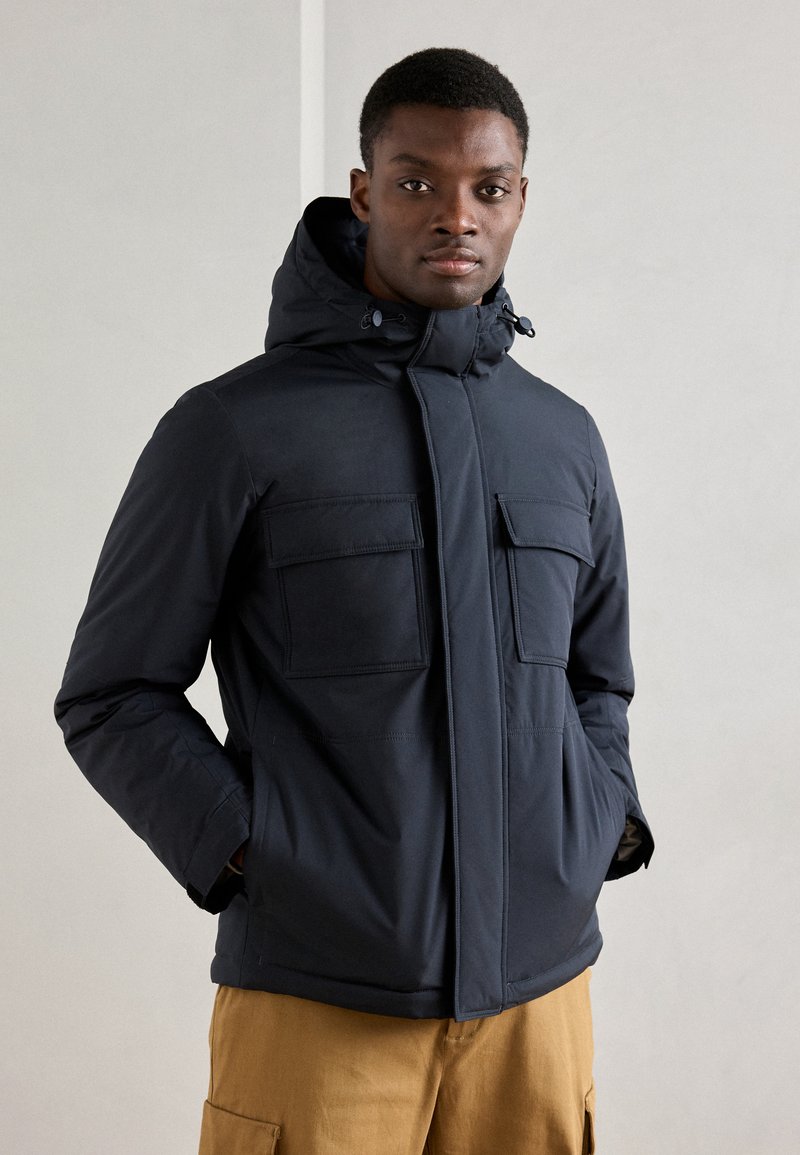 Casaco acolchoado azul-marinho com capuz, apresentando dois grandes bolsos frontais, um fecho éclair à frente e uma textura suave. Usado sobre calças bege.