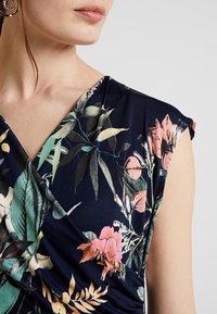 Granatowa sukienka w kwiaty z dekoltem kopertowym, krótkimi rękawami oraz żywymi zielonymi i różowymi wzorami kwiatowymi na gładkim materiale.