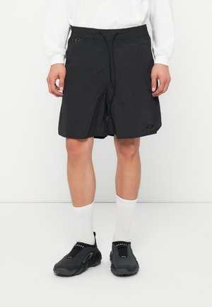Schwarze Shorts aus einem leichten, strukturierten Material, mit einem elastischen Bund, Kordelzug und Seitentasche. Getragen mit weißen Socken und schwarzen Schuhen.