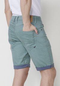 Shorts vert clair avec un ourlet roulé, détails de poche arrière et une étiquette en cuir sur la ceinture ; fabriqués en tissu texturé.