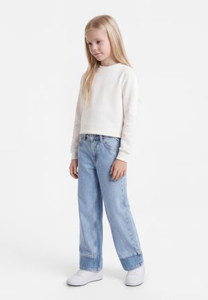 Mladé dievča s dlhými blond vlasmi, oblečené v bielej mikine, svetlomodrých širokých džínsach a bielych teniskách, stojace na jednoduchom pozadí.