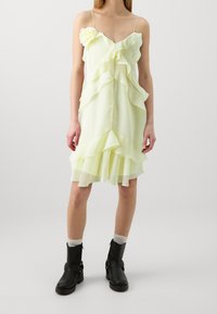 Světle žluté bezramenné šaty s volánovými vrstvami a květinovým akcentem na rameni. Nosené s černými kotníkovými botami a bílými ponožkami.