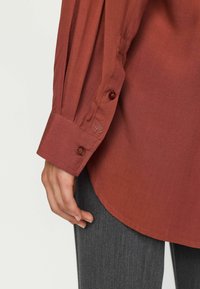 Detailní záběr ruky odpočívající na boku, oblečené v hnědorudém trikotu s dlouhými rukávy a tmavě šedých kalhotách.