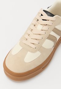 Beżowe zamszowe sneakersy z kremowymi akcentami, okrągłym noskiem i płaskimi sznurówkami. Posiadają gumową podeszwę i teksturowany wzór po bokach.