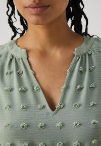 Απαλή πράσινη μπλούζα με V-λαιμό, που διαθέτει υφαντές ανθισμένες λεπτομέρειες και μαλακά συγκεντρωμένο λαιμόκοψη. Το ύφασμα είναι ελαφρύ και ελαφρώς τσαλακωμένο.