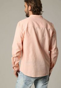 Licht perzikkleurig overhemd met lange mouwen en een button-down kraag, gestructureerde stof en zichtbare naden, gedragen over lichtblauwe jeans.