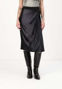 Black draped midi skirt with a smooth texture, featuring a side knot detail and a wide waistband, paired with knee-high glossy black boots.