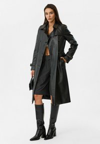 Zwarte leren trenchcoat met dubbelknopen, zijzakken en met riemen afgewerkte manchetten. Gecombineerd met hoge zwarte leren laarzen en grijze shorts.