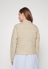 Beige denimitakki, jossa on muotoiltu malli, näkyvät ompeleet ja nappikoristeet. Takakuvassa näkyy rakenteellinen siluetti ja imarteleva leikkaus.