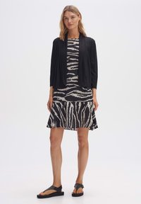 Zwarte wikkelcardigan over een zwart-wit gestreepte jurk met flared zoom. Model draagt zwarte platte sandalen; de gladde stoftexturen zijn zichtbaar.