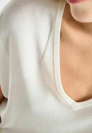 Nahaufnahme eines weißen V-Ausschnitt-Strickoberteils, das von einer Person getragen wird, mit sichtbarer Textur des Stoffes und Details am Halsausschnitt.