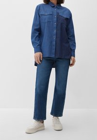 Kvinde iført tofarvet blå denimskjorte, mørkeblå jeans og hvide sneakers, der står mod en ensfarvet lys væg.