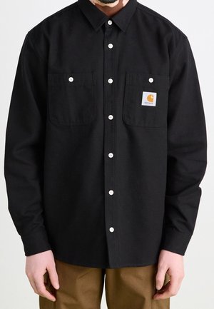 Hombre vistiendo una camisa negra con botones blancos y dos bolsillos en el pecho, uno de ellos con un pequeño parche con el logo de Carhartt.