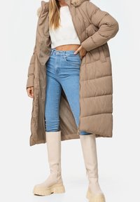 Długa beżowa pikowana kurtka z kapturem wykończonym sztucznym futrem, zapinana na zamek, z bocznymi kieszeniami, noszona na białym krótkim swetrze i niebieskich dżinsach; zestawiona z kremowymi kozakami sięgającymi do kolan.
