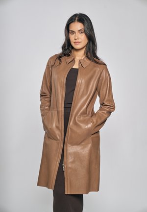 Femme portant un manteau en cuir marron mi-long sur un haut et une jupe foncés, debout les mains dans les poches du manteau, sur un fond uni.