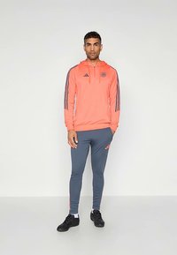 Férfi sportos öltözék: élénk korall színű kapucnis pulóver szürke csíkokkal és logóval, szürke, testhez simuló tréningnadrággal, rózsaszín részletekkel. Fekete sportcipő.