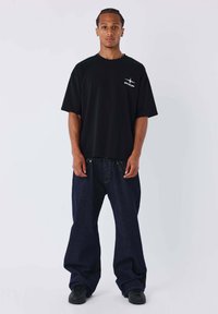 Maglietta nera in cotone con un piccolo logo bianco sul petto, abbinata a jeans larghi in denim blu scuro e sneaker nere. Design pulito e vestibilità perfetta.