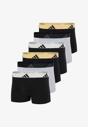 Muskulöser Mann steht barfuß da, trägt schwarze Boxershorts mit einem gelben Adidas-Bund und blickt zur Seite, während ein Arm auf dem anderen ruht.