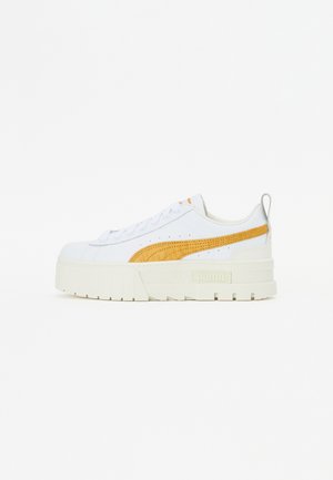 Białe sneakersy z teksturowanym żółtym paskiem, grubą białą podeszwą i szarym akcentem na pięcie. Zawierają wyraźne logo PUMA z boku.