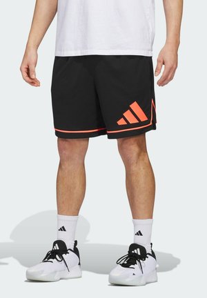 Mann trägt schwarze Sportshorts mit orangefarbener Einfassung und Logos, weiße Sneakersocken, weiße und schwarze Sportschuhe, steht vor hellem Hintergrund.
