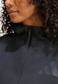 Svart zip-up jakke med høy krage, laget av glatt stoff. Har et teksturert logoakent og kontrasterende sømdetaljer.