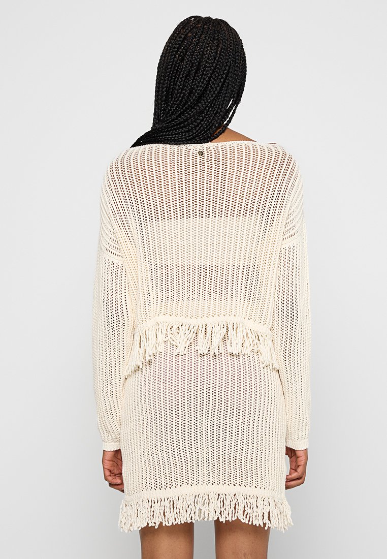 Abito maglione in bianco sporco, con lavorazione a maglia aperta, maniche lunghe e dettagli in frange all'orlo. Vestibilità rilassata con scollatura a V sulla schiena.