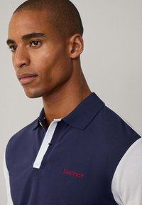 Marinblå pikétröja med vita ärmar, har en röd broderad logotyp "HACKETT" och en klassisk krage med knappstängning.
