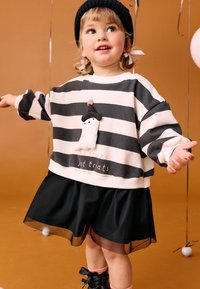 Felpa a righe in crema e navy, con una patch a forma di personaggio e testo. Gonna nera a strati con orlo trasparente; un cappello di maglia nera completa il look.