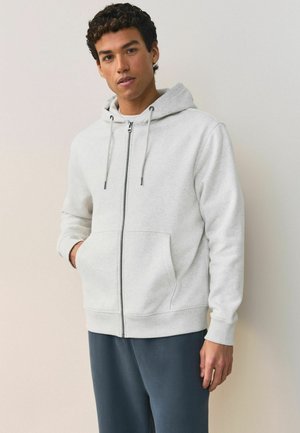 Junger Mann trägt eine hellgraue Kapuzenjacke mit Reißverschluss und hat die Hände in den vorderen Taschen, steht vor einer schlichten beigen Wand.