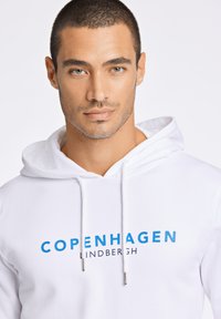 Sudadera blanca con un gráfico azul de "COPENHAGUE" y el texto "LINDBERGH". Cuerdas de ajuste con terminales plateados. Textura suave y lisa.