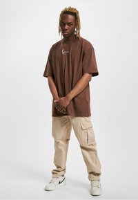 Camiseta marrón de gran tamaño con un logo blanco, combinada con pantalones cargo beige claros que tienen bolsillos laterales. El modelo lleva zapatillas deportivas blancas.