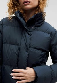 Veste rembourrée bleu marine foncé avec un col haut, un motif matelassé et un bouton sur le côté. Tissu doux et brillant avec une silhouette ajustée.